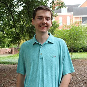 A young man with a mustache smiles while wearing a light blue polo shirt in a park setting.