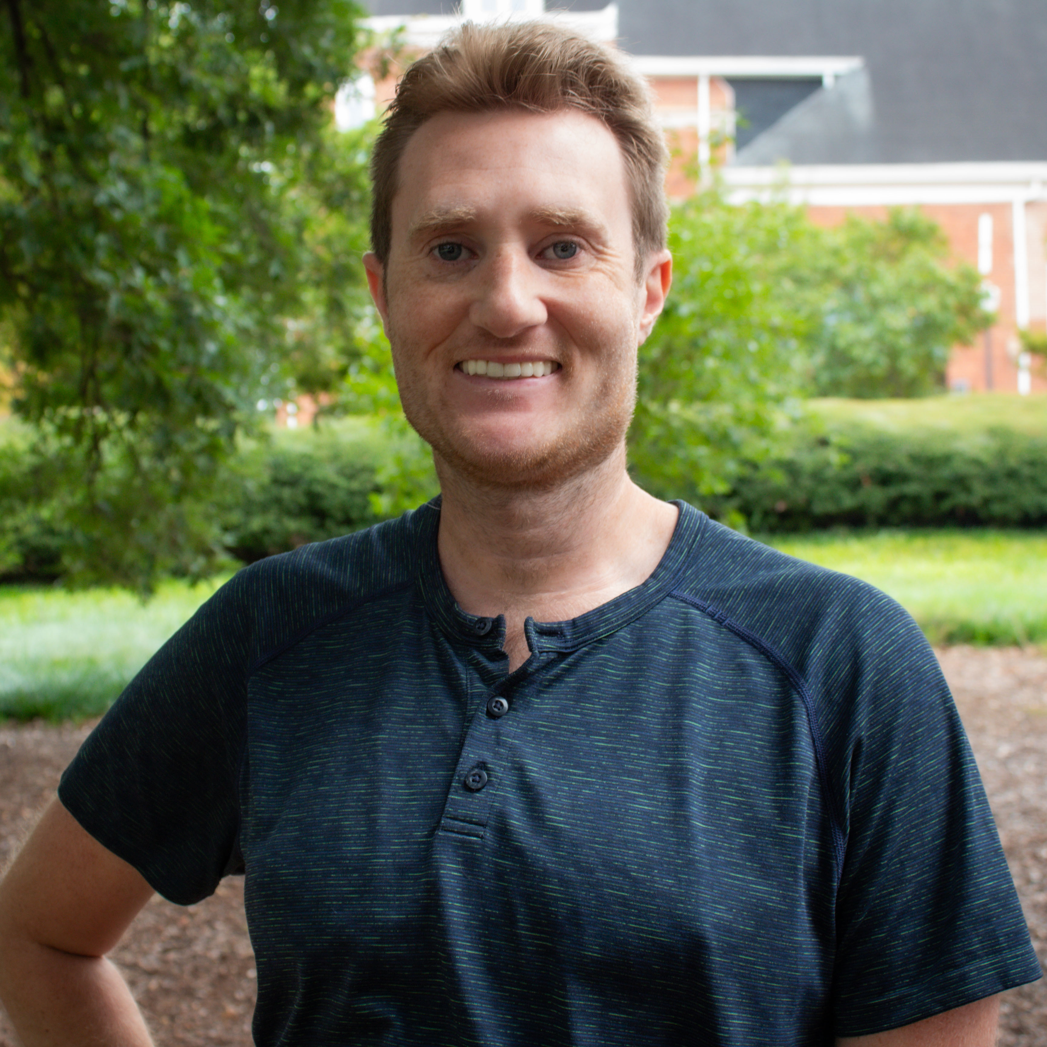A smiling man standing outdoors in a casual navy blue shirt, surrounded by greenery and a blurred background of a building.