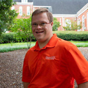 A smiling individual in an orange polo shirt stands in a garden setting with greenery and brick buildings in the background.