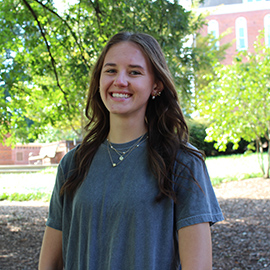 Smiling young woman standing outdoors under trees with a campus building in the background.