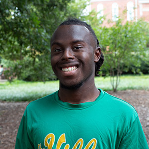 Smiling young man wearing a green shirt, standing outdoors with greenery in the background.