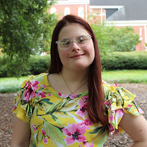 A young woman with long red hair and glasses smiles while wearing a colorful floral blouse in an outdoor setting.