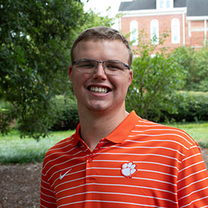 A smiling young man wearing an orange and white striped polo shirt with a Clemson University logo, standing outdoors in a green area.