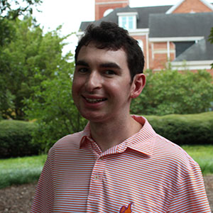 A young man smiles while sitting outdoors in a garden setting.