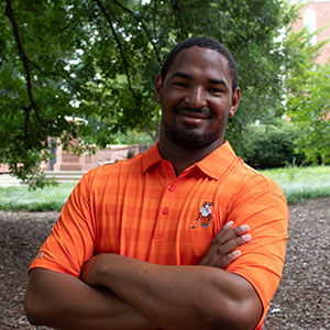 A smiling man in an orange polo shirt, standing outdoors with arms crossed and trees in the background.