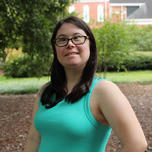 A smiling woman with glasses stands outdoors, wearing a teal tank top.