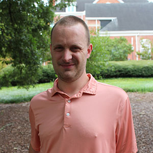 A smiling man in a pink polo shirt stands outdoors in a grassy area with trees and a building in the background.