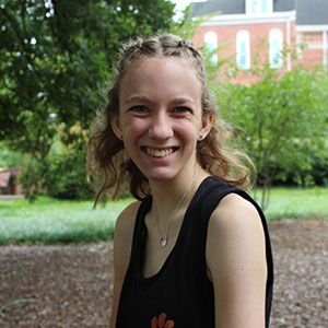 A young woman smiles while sitting outdoors in a garden setting.