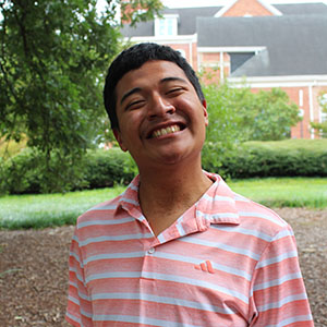 A young man with a big smile stands outdoors in a casual striped pink shirt, surrounded by greenery.