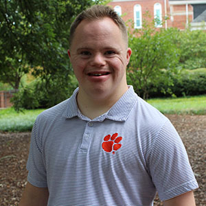 A young man smiles while sitting outdoors in a garden setting.