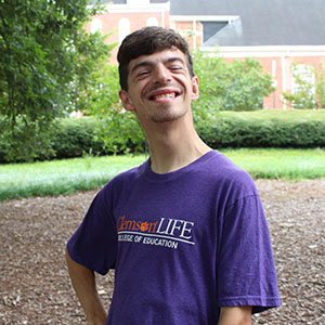 Smiling student wearing a purple Clemson LIFE T-shirt, posing outdoors with greenery in the background.