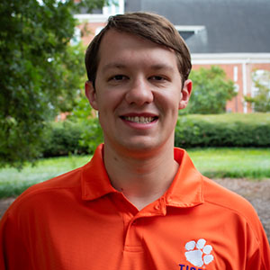 Smiling man wearing an orange polo shirt with a Clemson University logo, standing outdoors in a lush green setting.