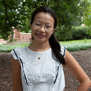 A young woman with glasses smiles confidently while standing outdoors in a park-like setting.