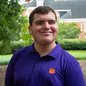 Young man wearing a purple polo shirt with a Clemson University logo, smiling outdoors in a park-like setting.