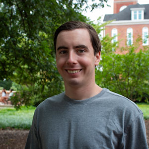 A young man smiles while sitting outdoors in a garden setting.