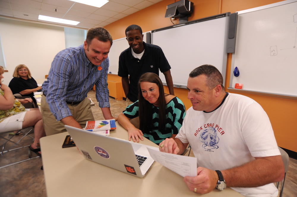 A group of four individuals collaborating around a laptop in a classroom setting, focused on a document and discussing ideas.