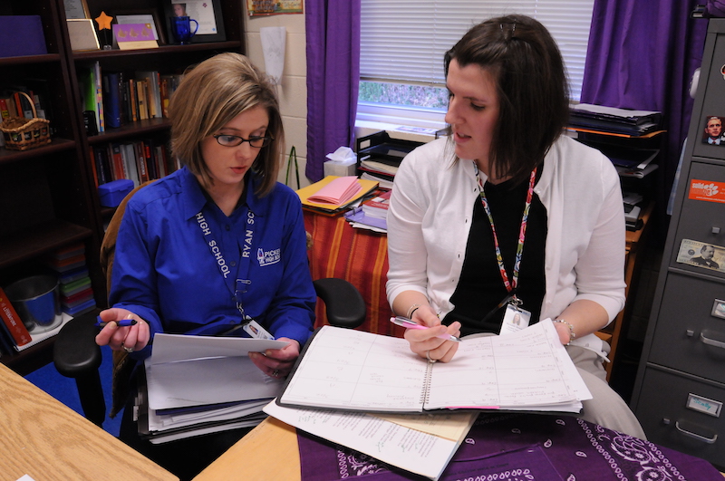 Teachers collaborating in a classroom, with one reviewing documents and the other making notes in a planner.