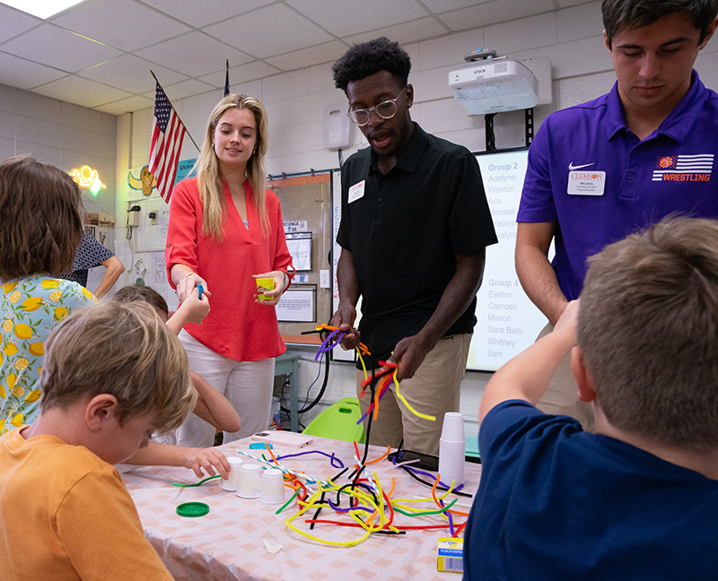 Three adult volunteers help children with a hands-on craft at a classroom table covered with colorful pipe cleaners and supplies.