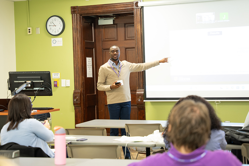 Graduate student at the front of a classroom pointing at a projected slide while students watch from their desks.