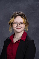 Portrait of a young woman with glasses and wavy hair, wearing a black blazer over a red shirt.