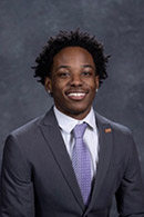 Professional portrait of a young man in a suit and tie, smiling confidently.