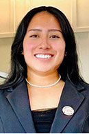 A smiling young woman wearing a black blazer and pearl necklace, standing in a well-lit environment.