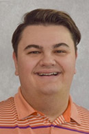 Headshot of a young man with short hair, smiling and wearing an orange striped polo shirt.
