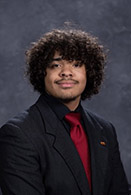 A professional headshot of a young man with curly hair, wearing a black suit and a red tie, set against a gray background.