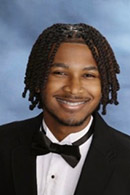 High school senior portrait featuring a young man with braided hair, wearing a tuxedo and bow tie, smiling confidently against a soft blue background.