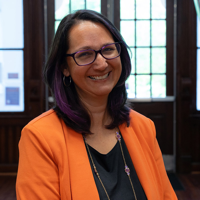 Professional portrait of a smiling woman with purple highlights, wearing an orange blazer and glasses.