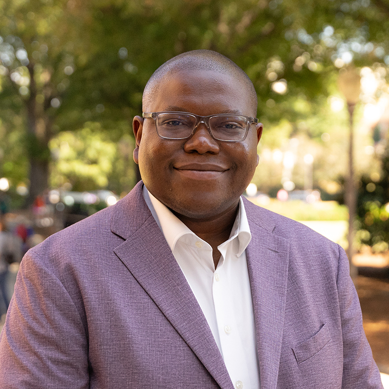 A smiling man in a lavender blazer and white shirt, standing outdoors with trees in the background.