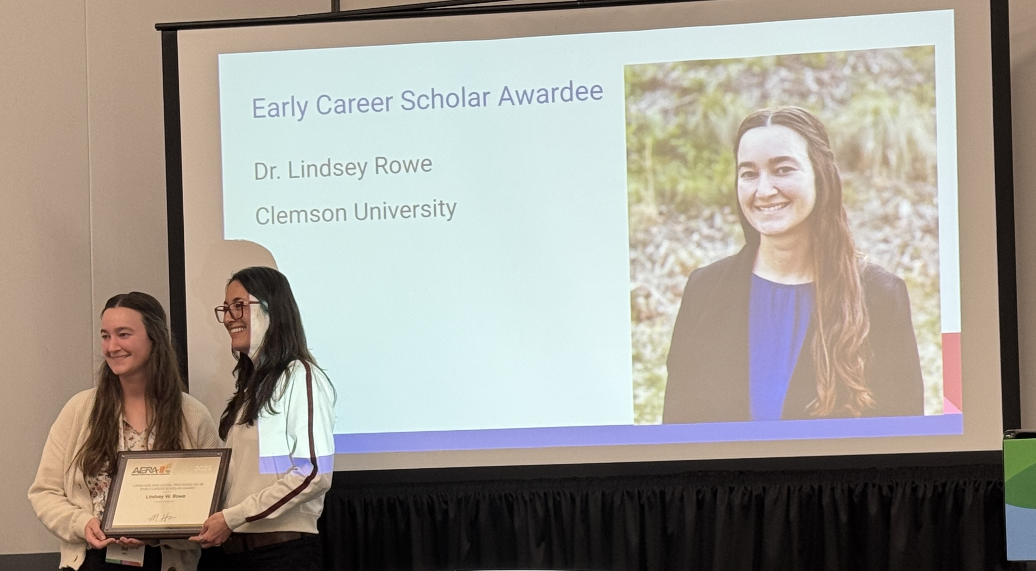 Two women are standing together, one holding a certificate, with a large screen behind them displaying a photo of Dr. Lindsey Rowe alongside her name and affiliation with Clemson University.