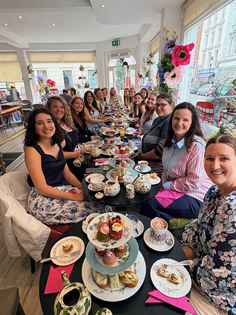 Large group seated along a long table enjoying afternoon tea with tiered trays of pastries and teapots in a bright, flower-decorated cafe.