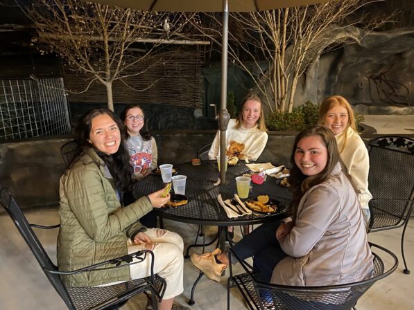 Five friends smiling around an outdoor patio table with drinks and food under a large umbrella at night.