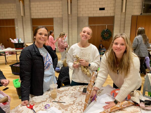 Three young women smile while wrapping gifts at a table covered with wrapping paper and supplies in a gym or community hall.