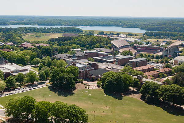 Aerial view of campus with Bowman field in the center and Tillman hall behind it. The football stadium is off to right with the lake and trees on the horizon. 