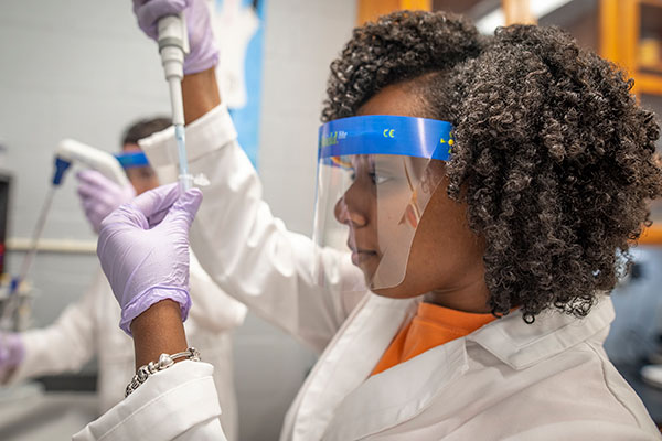 Woman in a white lab coat and latex gloves is holing a dropper in a tube.