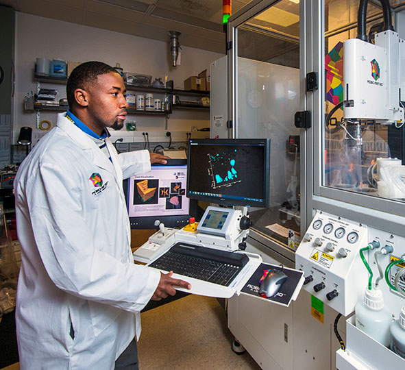 Male student in white lab coat stands in front of a keyboard and computer screen controlling a robot behind a glass wall. 