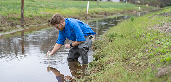 Male student wearing waterproof waders retrieves a water sample from a stream.