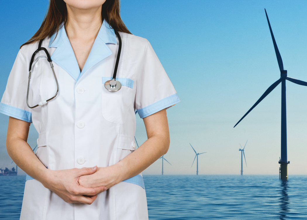 A woman in a nurse uniform and stethoscope stands with hands clasped with an offshore wind farm in the background