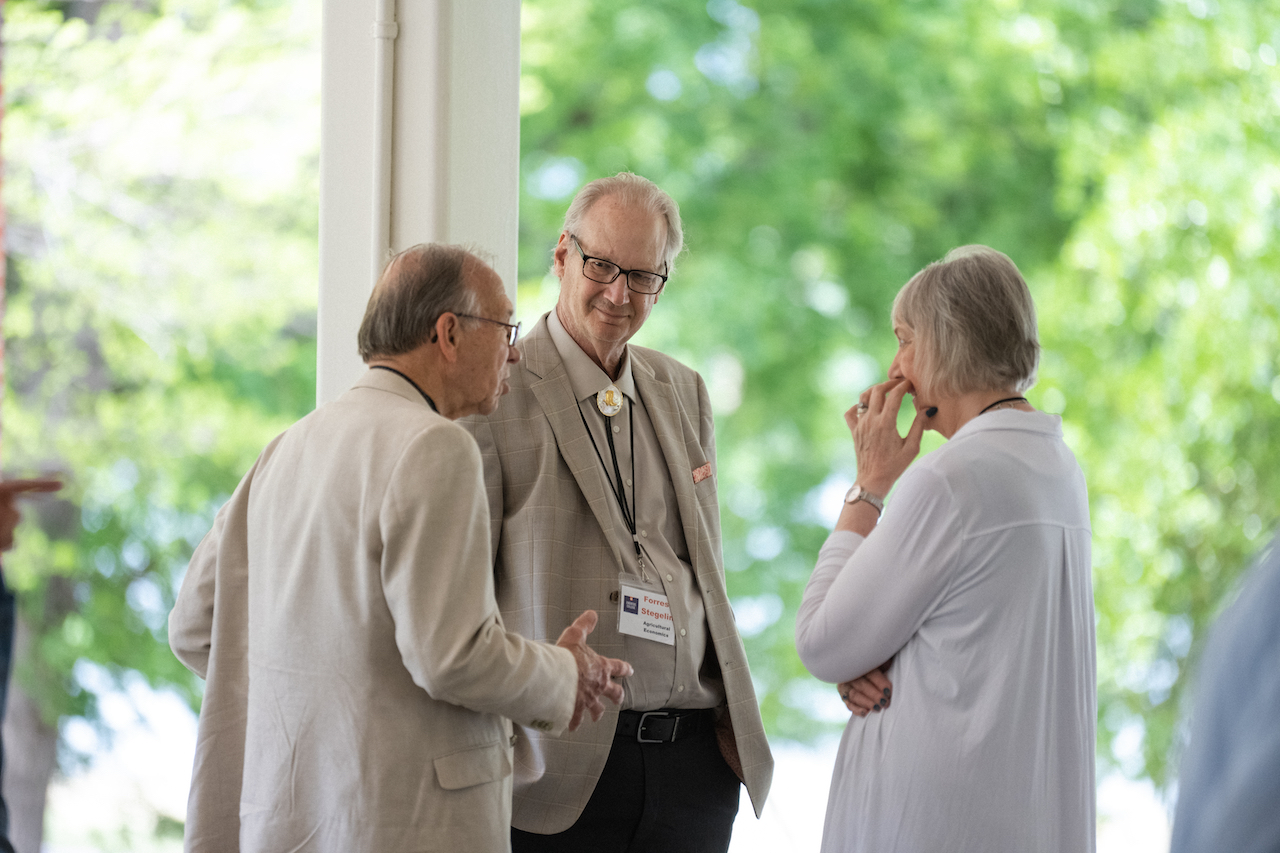 two men and a woman stand talking in front of a wooded background at an Emeritus College event