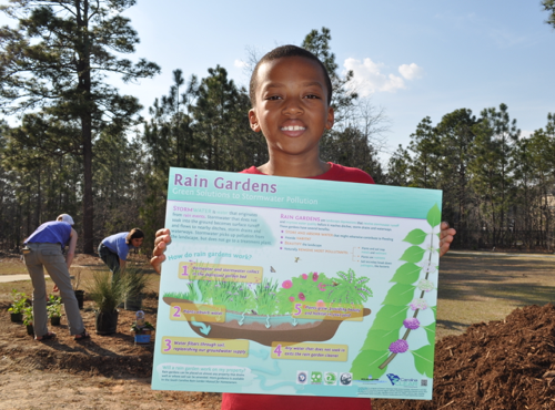 young boy standing outside on a sunny day holding a rain garden banner