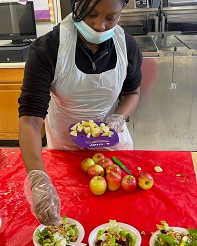 Woman wearing a face mask and plastic apron stands in front of red table placing cut up apples onto three salad plates