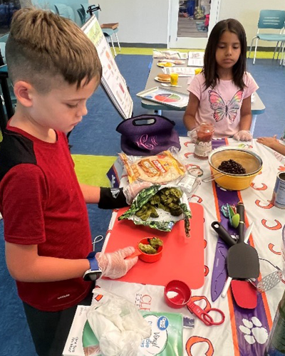 Child standing in front of red table scoops vegetables from bag into measuring cup