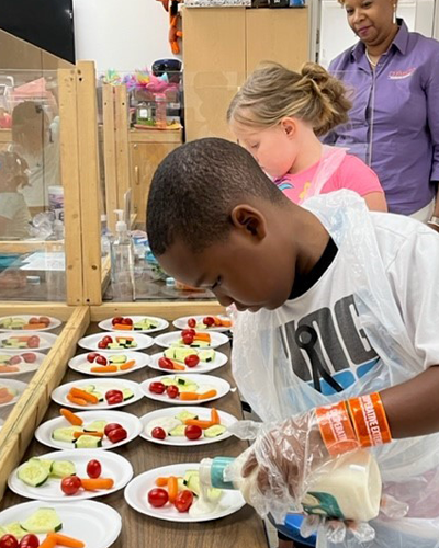 Child wearing plastic apron and food gloves pours ranch dressing onto different snack plates filled with a variety of vegetables