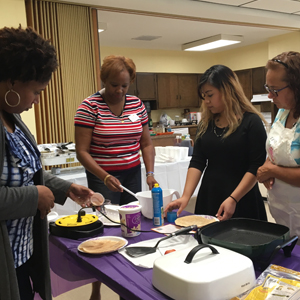 Four women standing around a purple table with different cooking utensils and ingredients