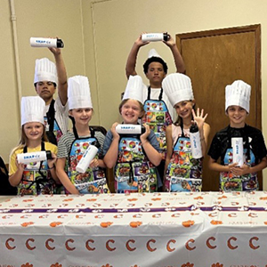 A group of students wearing chef hats stand in front of table with a Clemson table cloth holding water bottles