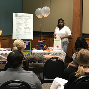 Women stands in front of group beside a table with balloons and different posters