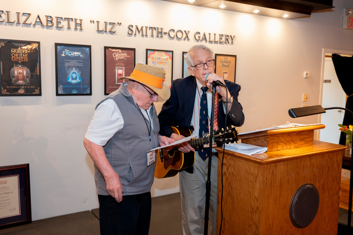 man holding a guitar speaks into a microphone as another man standing near reads from a page