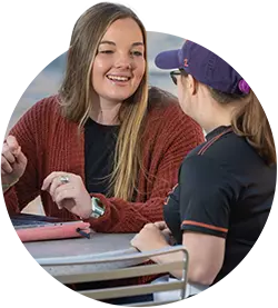 A female student sits at a table talking with another student
